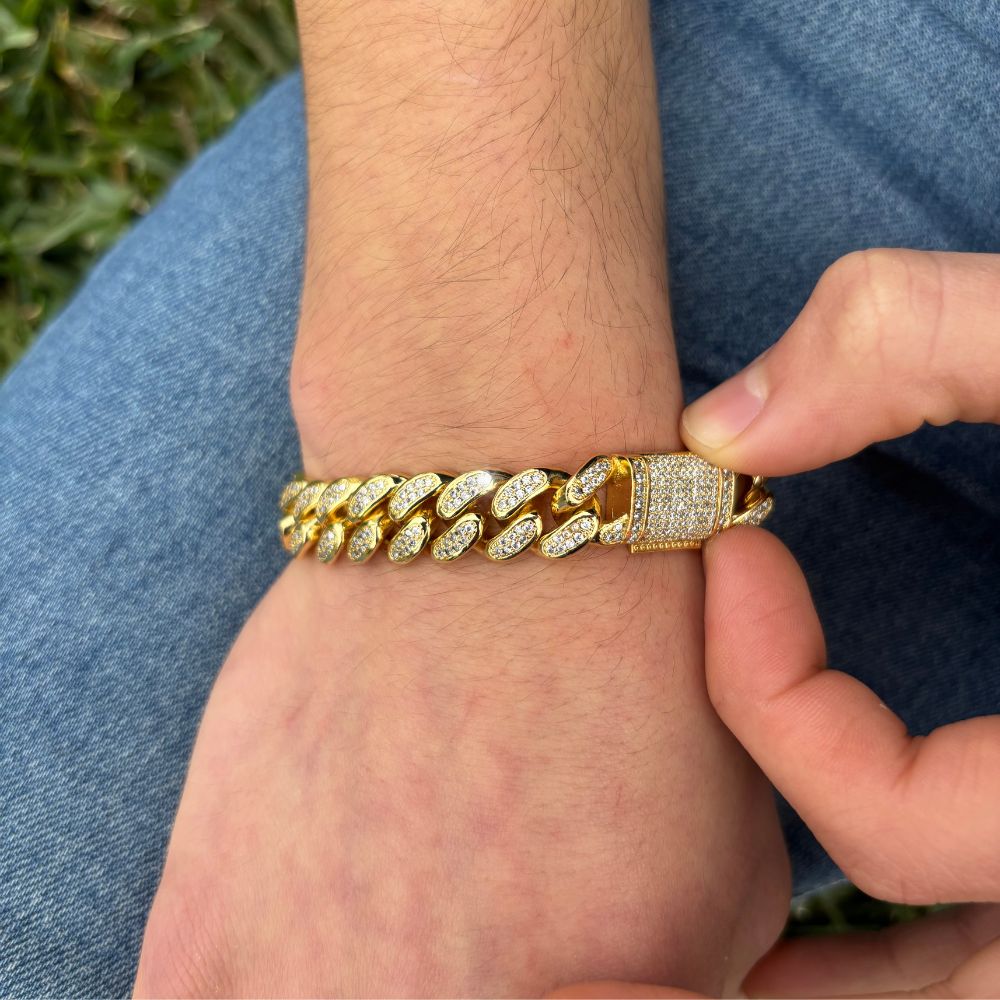 Modelo a mostrar a sua pulseira 12mm Iced Miami Cuban Link em ouro, com elos largos e incrustações de pedras brilhantes que criam um efeito luxuoso. A pulseira imponente destaca-se no pulso do modelo, oferecendo um visual sofisticado e ousado, perfeito para quem quer um estilo moderno e de alto impacto.
